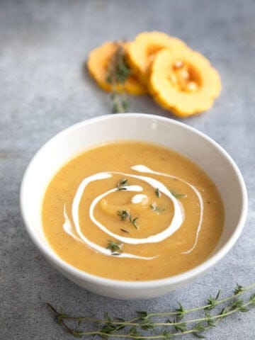 A white bowl filled with Roasted Delicata Squash Soup on a gray table, with rings of delicata squash in the background.