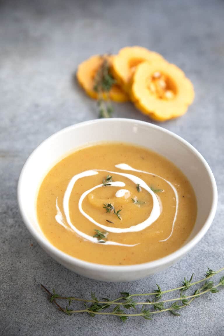 A white bowl filled with Roasted Delicata Squash Soup on a gray table, with rings of delicata squash in the background.