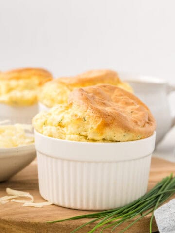 A Keto Cheese Soufflé in a white ramekin on a wooden cutting board, with chives.