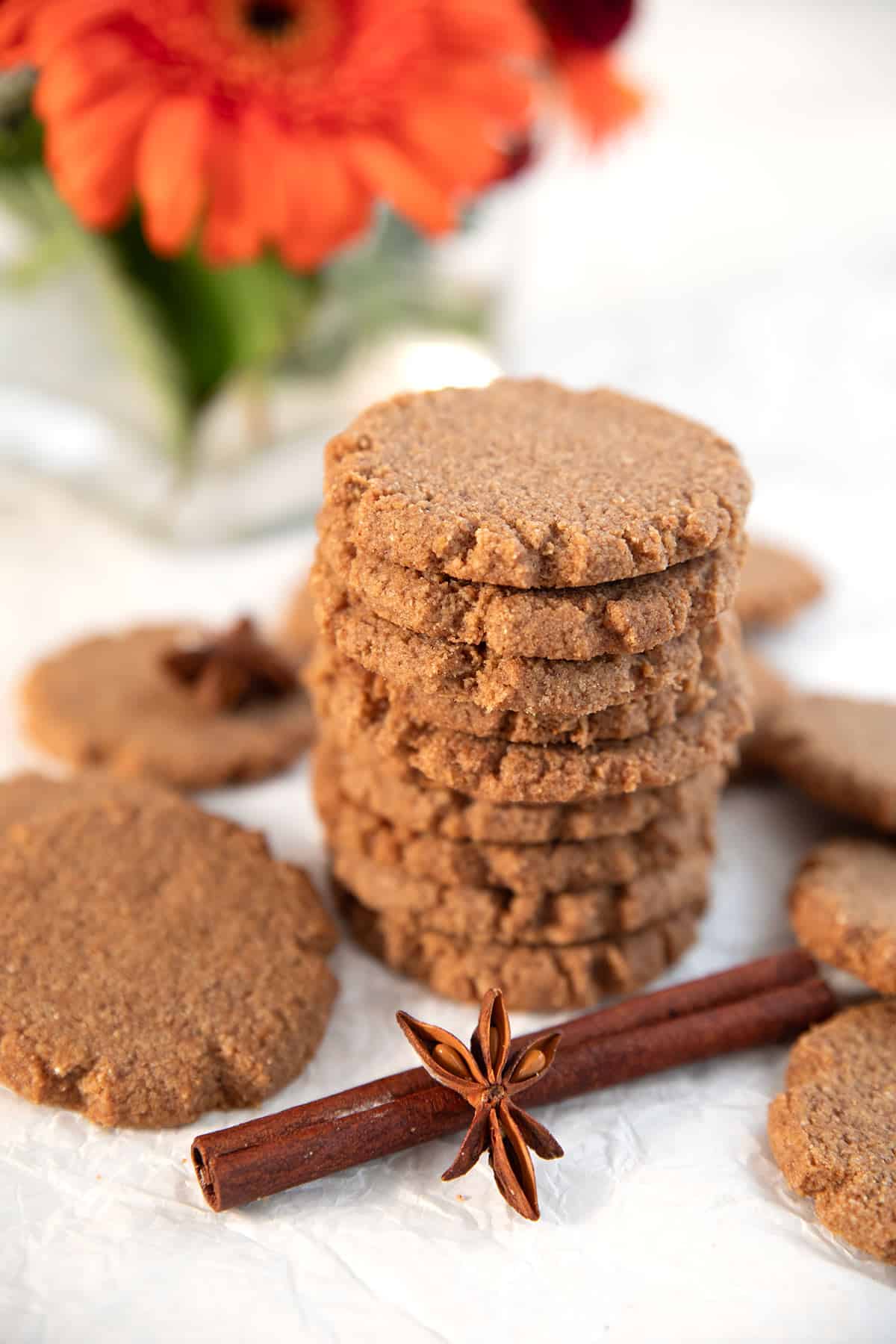 A stack of Keto Gingersnap Cookies with cinnamon and star anise, and orange Gerbera daisies in the background.