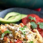 Titled image of a serving of chorizo and eggs on a black plate with avocado and tomatoes.