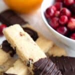 Titled image of Keto Cranberry Orange Shortbread Sticks piled up on white parchment with a bowl of cranberries and an orange in the background.