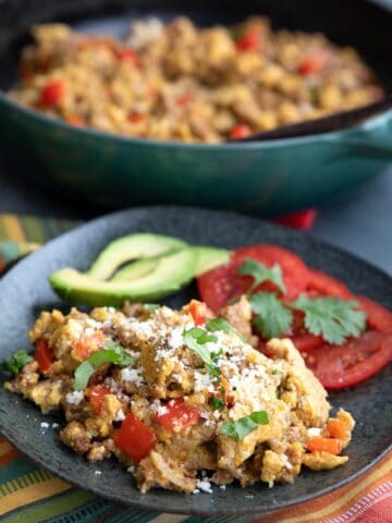 Keto Chorizo and Eggs on a black plate with tomatoes and avocado, with a pan of more eggs in the background.