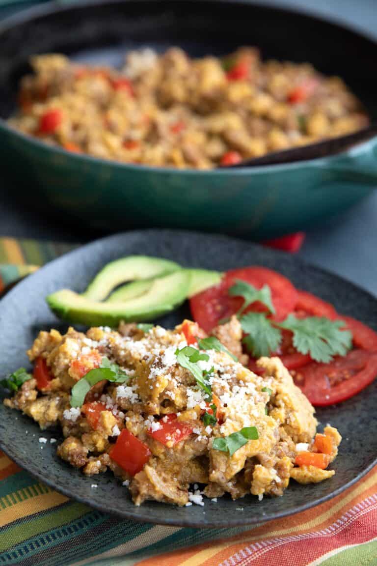 Keto Chorizo and Eggs on a black plate with tomatoes and avocado, with a pan of more eggs in the background.