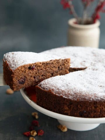 A slice of keto fruitcake being lifted away from the rest of the cake on a white cake stand.