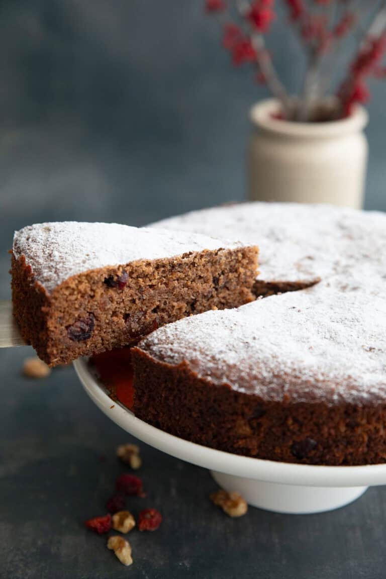 A slice of keto fruitcake being lifted away from the rest of the cake on a white cake stand.