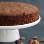 Titled image of a keto holiday fruit cake on a white cake stand, with dried cranberries and chopped walnuts all around.