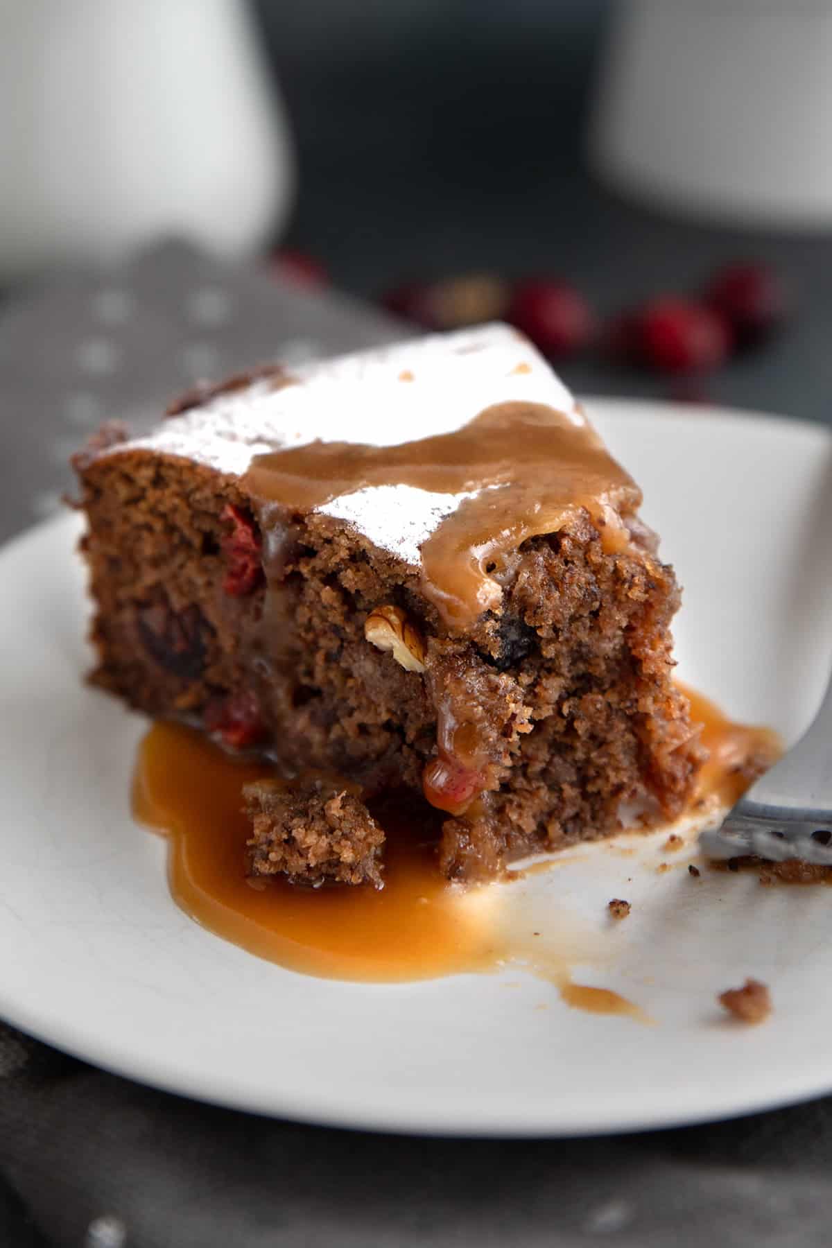 A slice of fruitcake drizzled with caramel sauce on a white plate, with several forkfuls taken out of it.