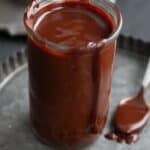 Titled image of Keto Hot Fudge Sauce in a glass jar on a metal tray, with two pieces of chocolate in front of it.