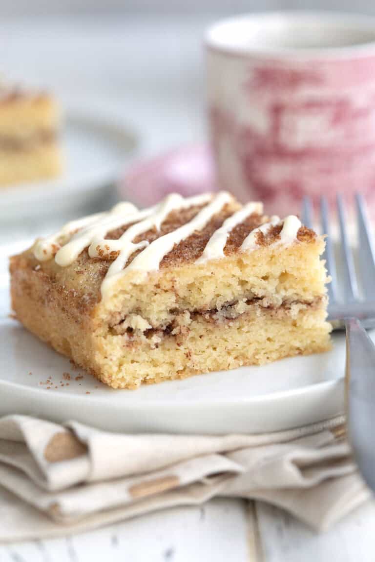 Close up shot of a slice of Keto Cinnamon Roll Coffee Cake on a white plate with a patterned red coffee cup in the background.