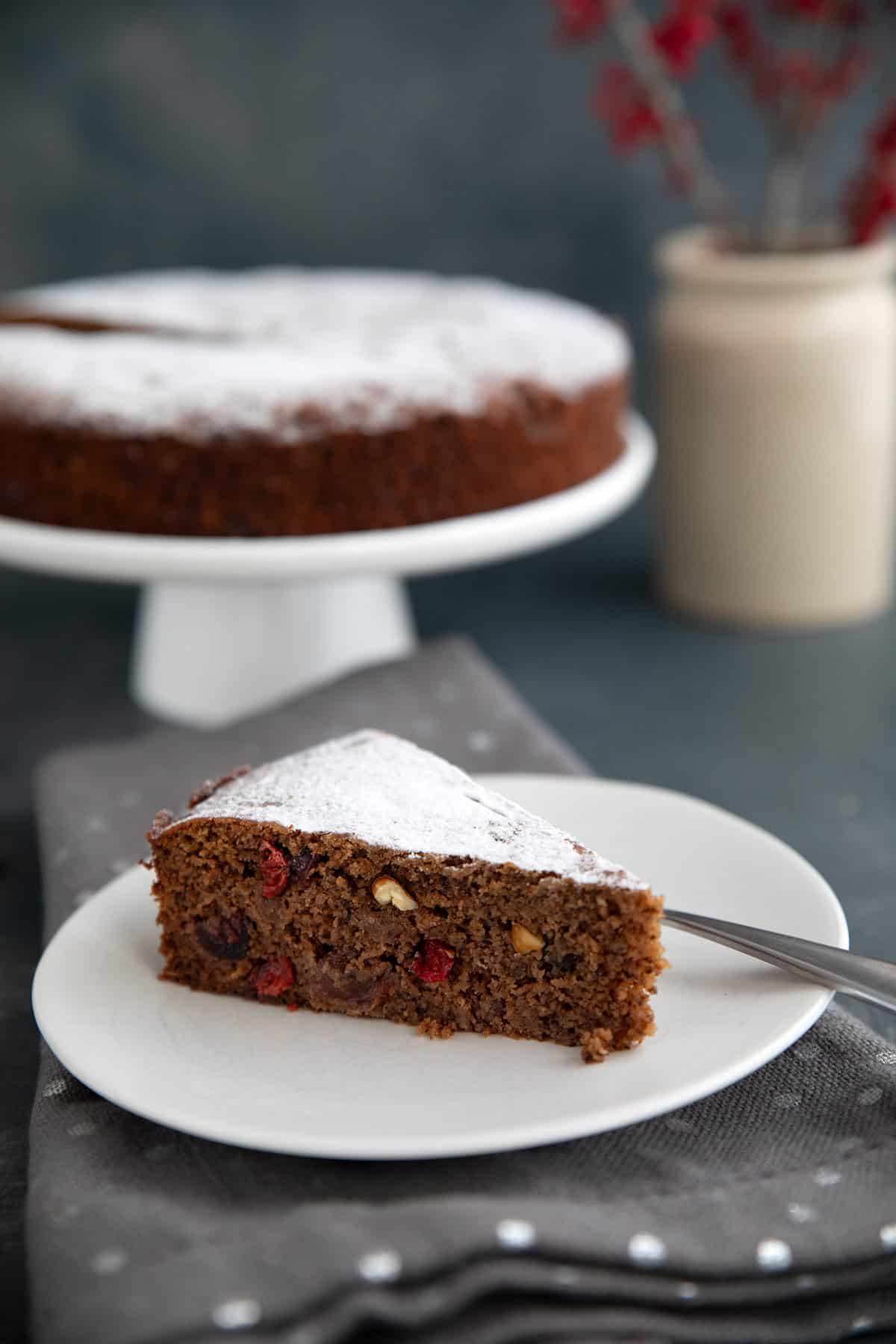 A slice of keto fruitcake on a white plate over a gray napkin, with the rest of the cake on a white cake stand in the background.