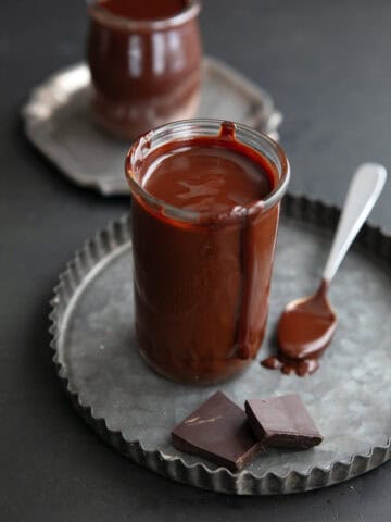 Two glass jars filled with keto hot fudge sauce sit on metal plates, the front one with a drip down the jar and two pieces of chocolate in front of it.