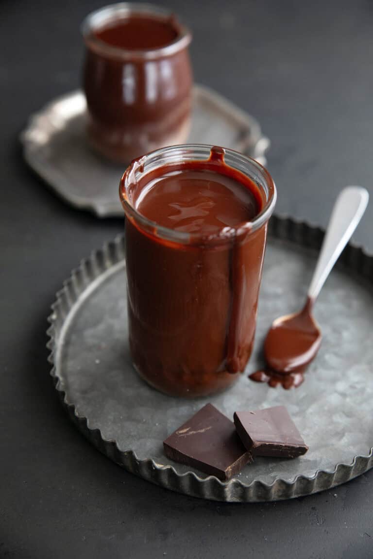 Two glass jars filled with keto hot fudge sauce sit on metal plates, the front one with a drip down the jar and two pieces of chocolate in front of it.