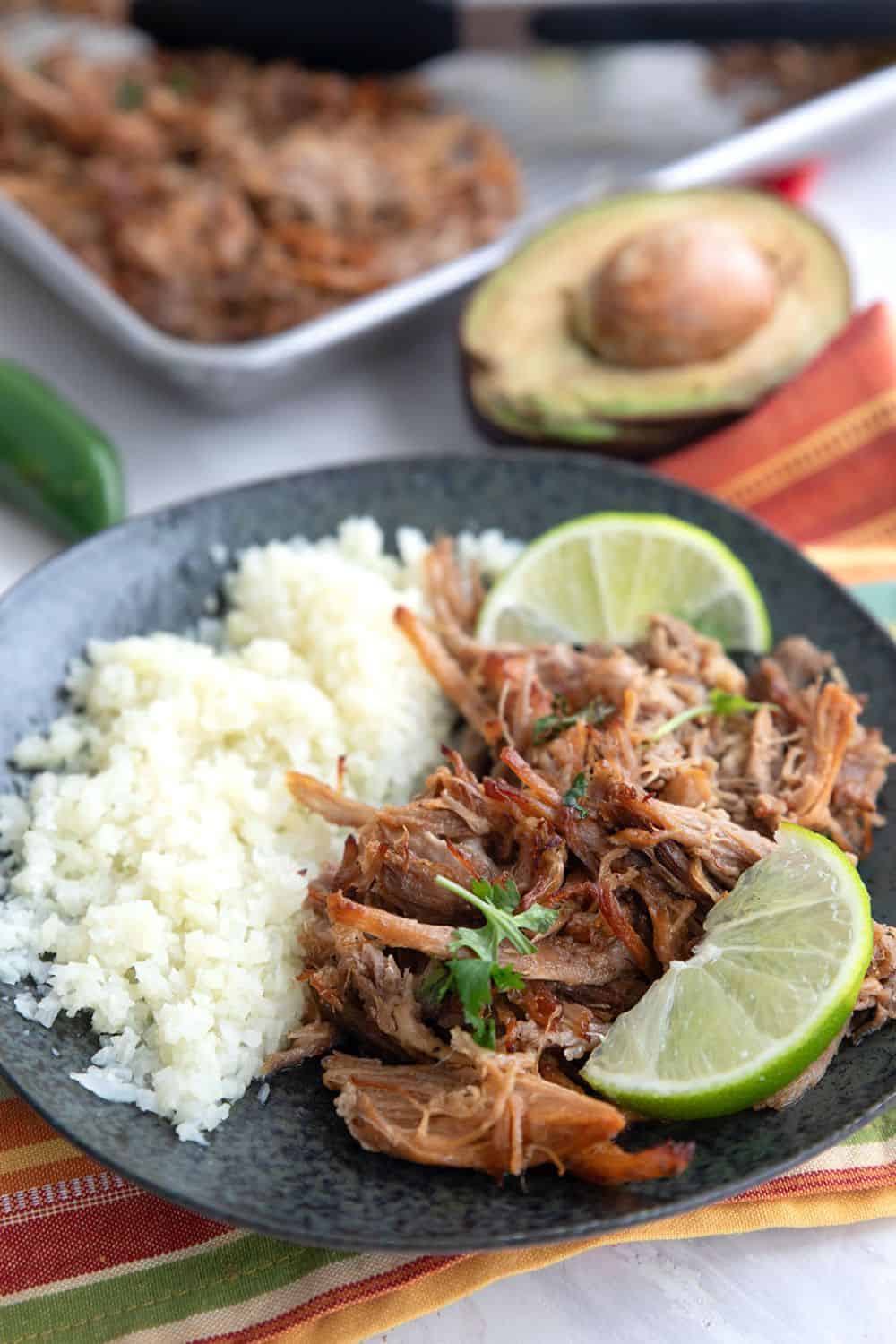 Crispy carnitas on a black plate with cauliflower rice and lime slices, set over a striped napkin.