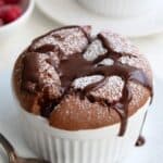 Titled image of a Keto Chocolate Soufflé in a white ramekin with powdered sweetener and chocolate sauce drizzled overtop.