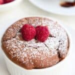 Close up shot of Keto Chocolate Soufflé in a small white ramekin with powdered sweetener and two raspberries on top.