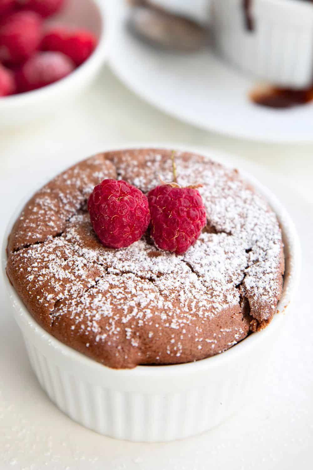 Close up shot of Keto Chocolate Soufflé in a small white ramekin with powdered sweetener and two raspberries on top.