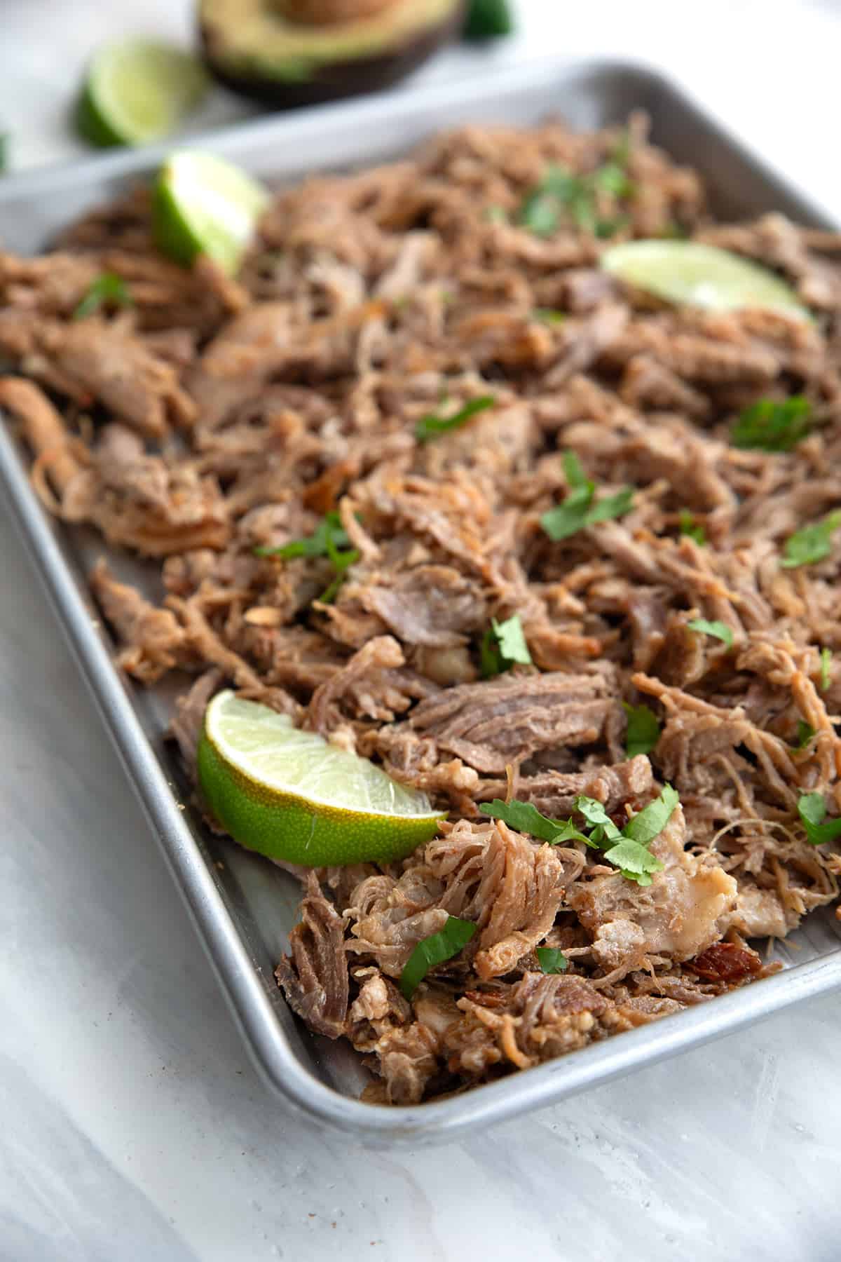 Crispy carnitas spread out on a metal baking pan on a white marble tabletop.