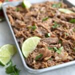 Crispy keto carnitas spread out on a baking pan with slices of lime and cilantro.