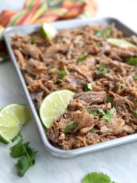 Crispy keto carnitas spread out on a baking pan with slices of lime and cilantro.