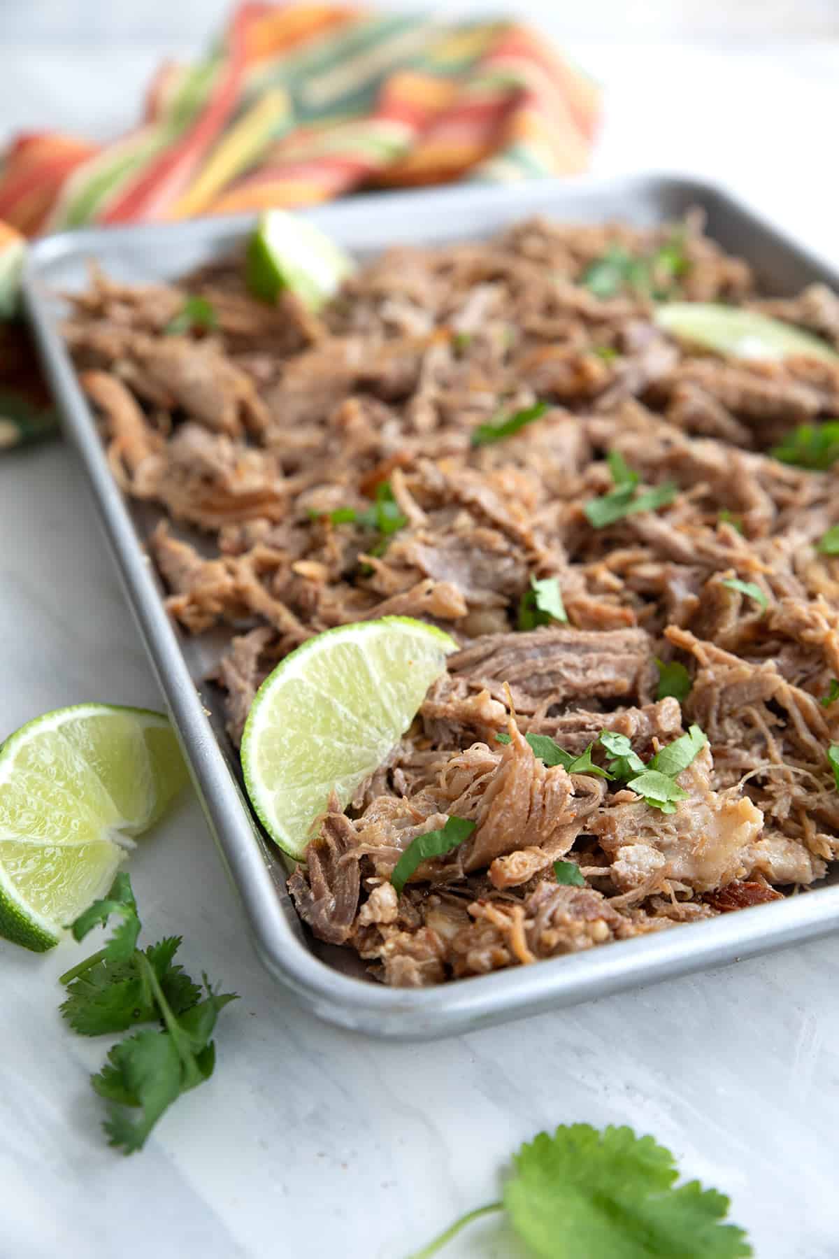 Crispy keto carnitas spread out on a baking pan with slices of lime and cilantro.