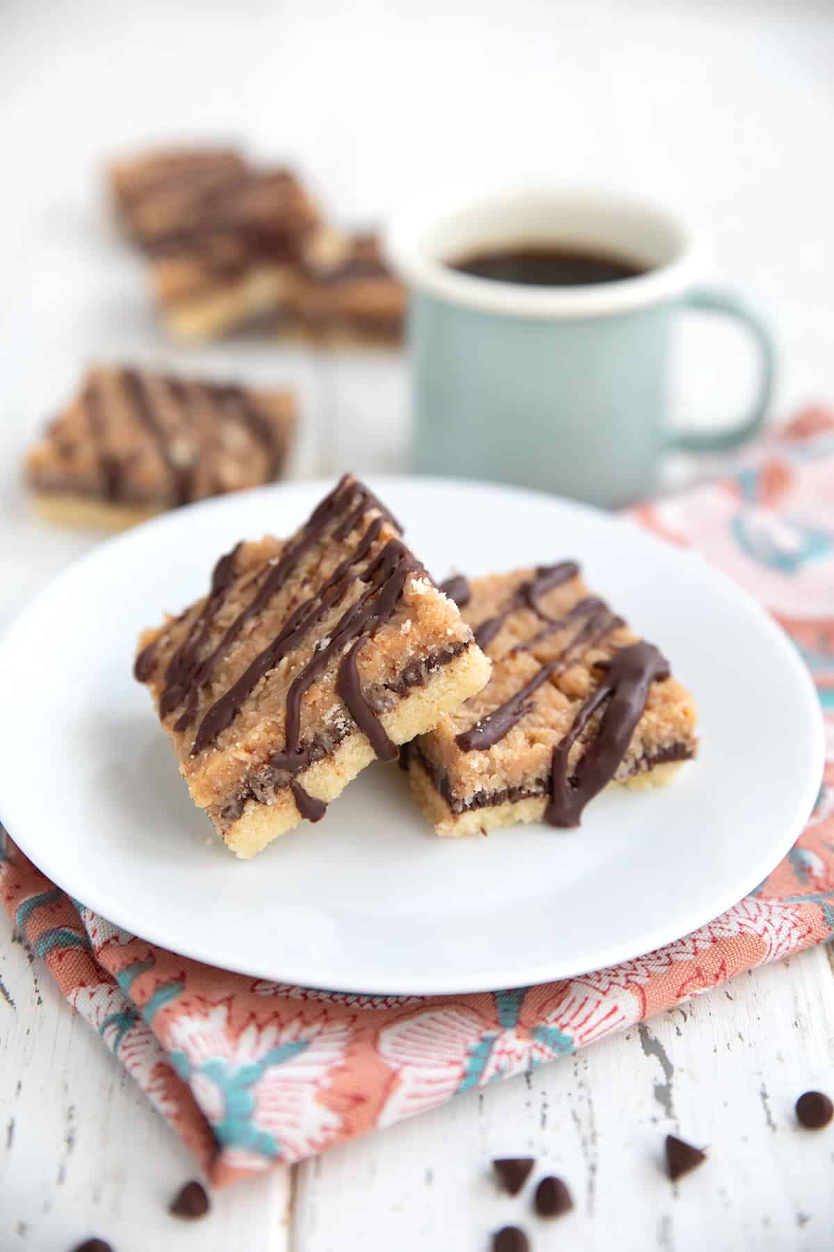 Two Keto Samoa Bars on a white plate over a pink patterned napkin, with a cup of coffee in the background.