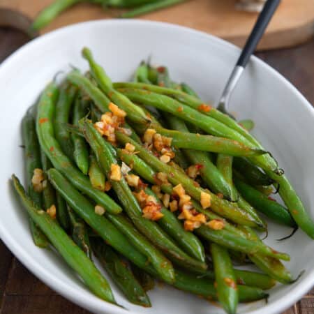 A white bowl filled with sautéed green beans with garlic and chili sauce on a brown wooden table.