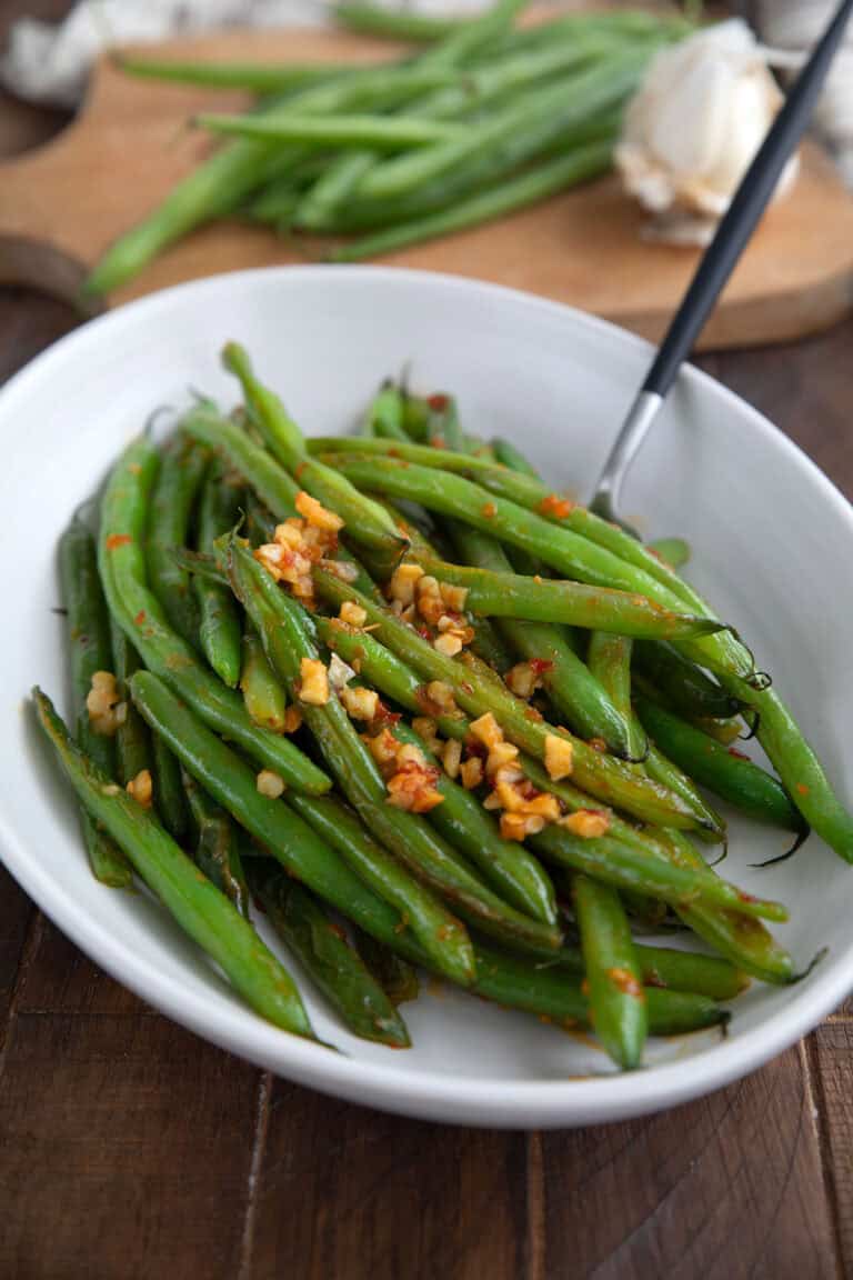 A white bowl filled with sautéed green beans with garlic and chili sauce on a brown wooden table.