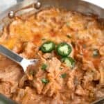 A metal serving spoon digging into a pan filled with Chicken Enchilada Skillet.