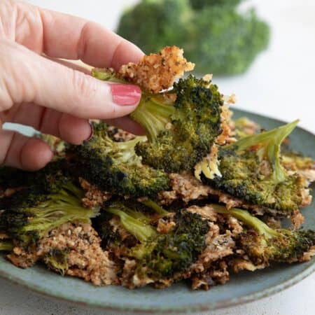 A hand holding up a piece of smashed broccoli above a bowl of more broccoli.