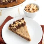 A slice of Keto Peanut Butter Cookie Cake on a white plate over a dark orange napkin, with a bowl of peanuts in the background.