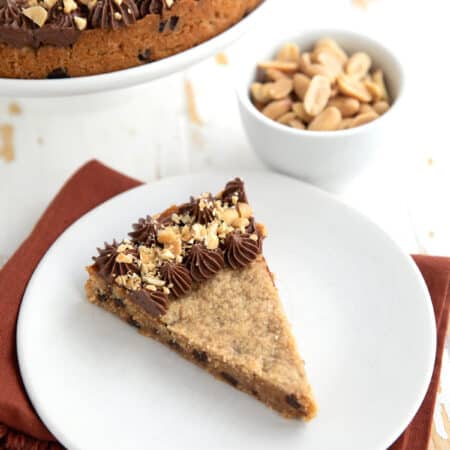 A slice of Keto Peanut Butter Cookie Cake on a white plate over a dark orange napkin, with a bowl of peanuts in the background.