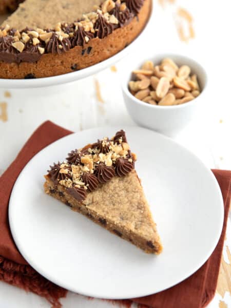 A slice of Keto Peanut Butter Cookie Cake on a white plate over a dark orange napkin, with a bowl of peanuts in the background.