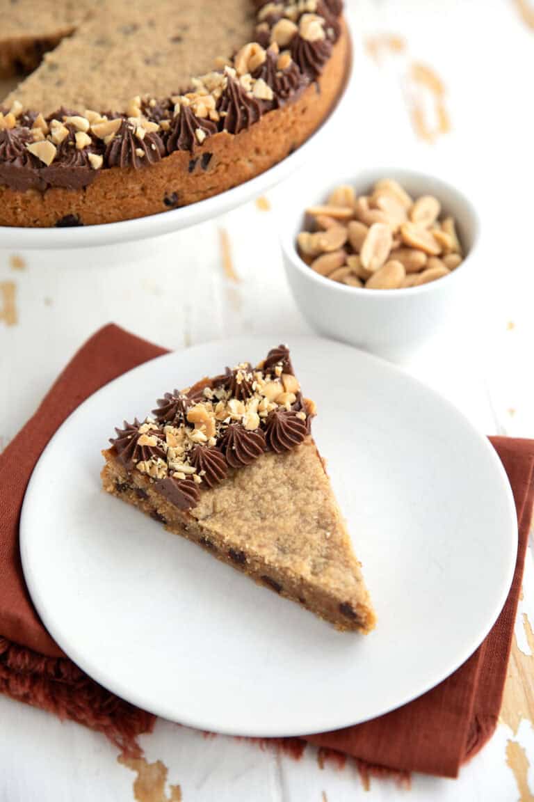 A slice of Keto Peanut Butter Cookie Cake on a white plate over a dark orange napkin, with a bowl of peanuts in the background.