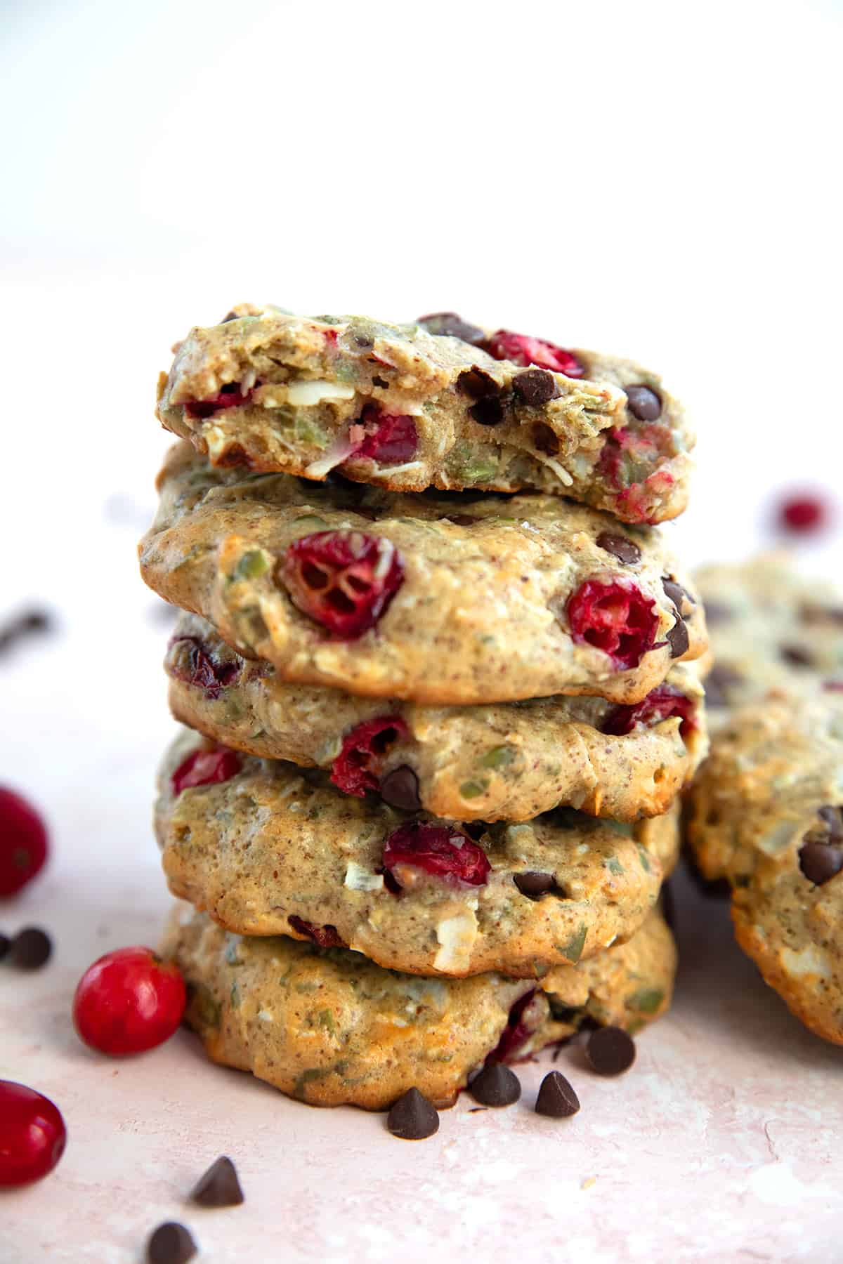 A stack of Protein Breakfast Cookies on a pink-hued table with fresh cranberries and chocolate chips strewn around.