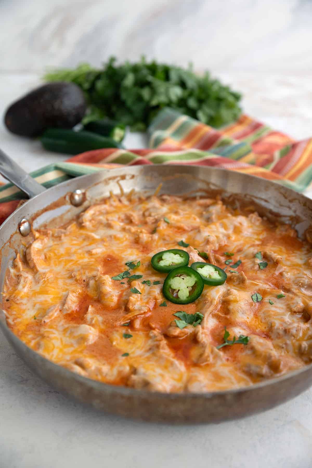 A metal pan filled with Chicken Enchilada Skillet on a white table, with cilantro, avocado, and jalapeño in the background.