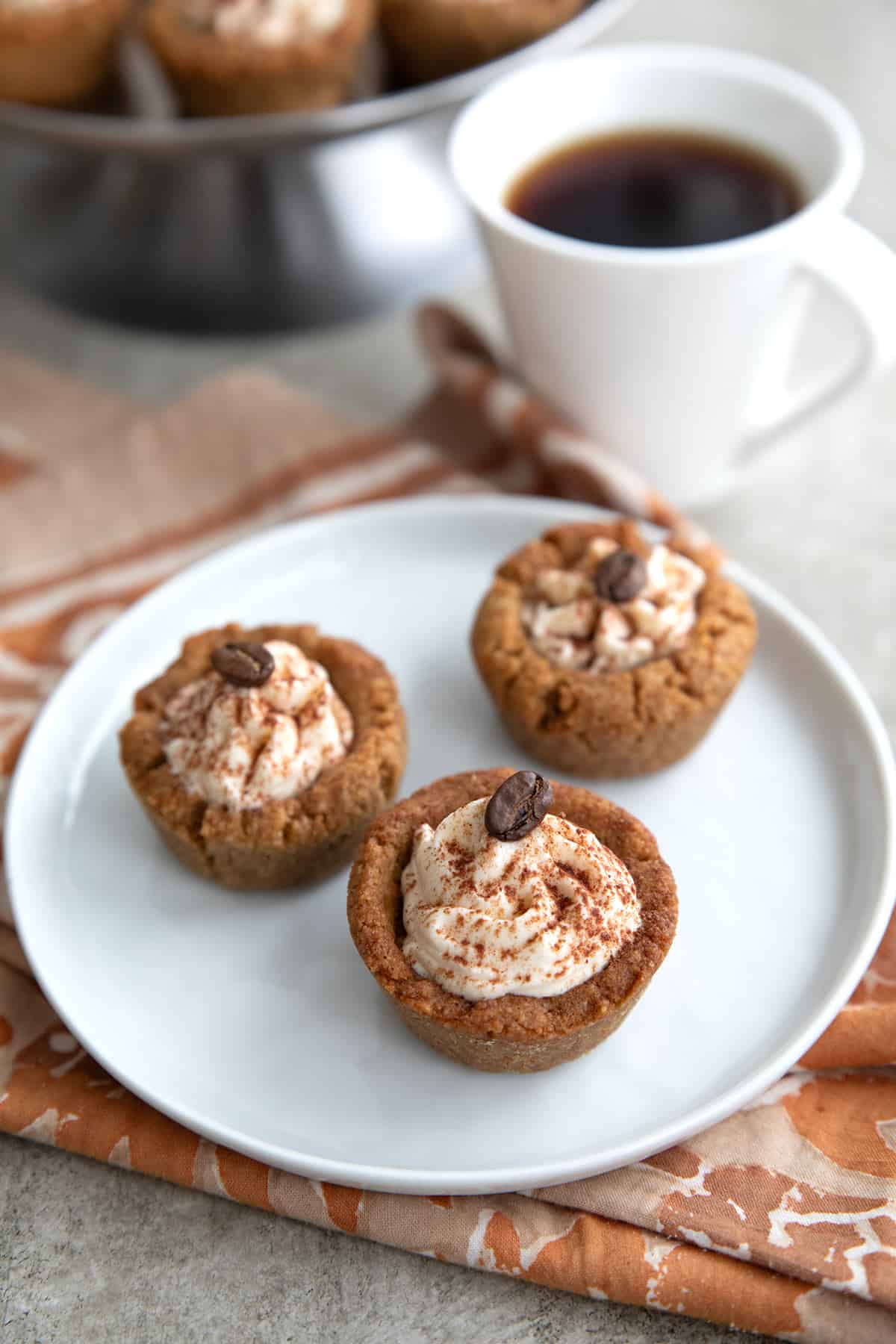 Three Keto Tiramisu Cookie Cups on a white pate with a cup of coffee in the background.