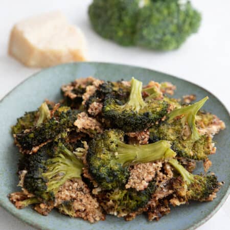 Crispy smashed broccoli piled up on a rustic green plate with parmesan and broccoli florets in the background.