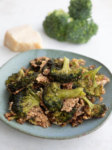 Crispy smashed broccoli piled up on a rustic green plate with parmesan and broccoli florets in the background.