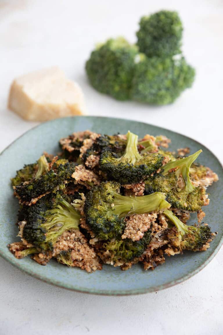 Crispy smashed broccoli piled up on a rustic green plate with parmesan and broccoli florets in the background.