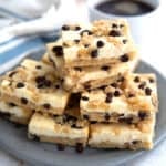 Keto Chocolate Chip Cookie Cheesecake Bars piled up on a plate with a cup of coffee in the background.