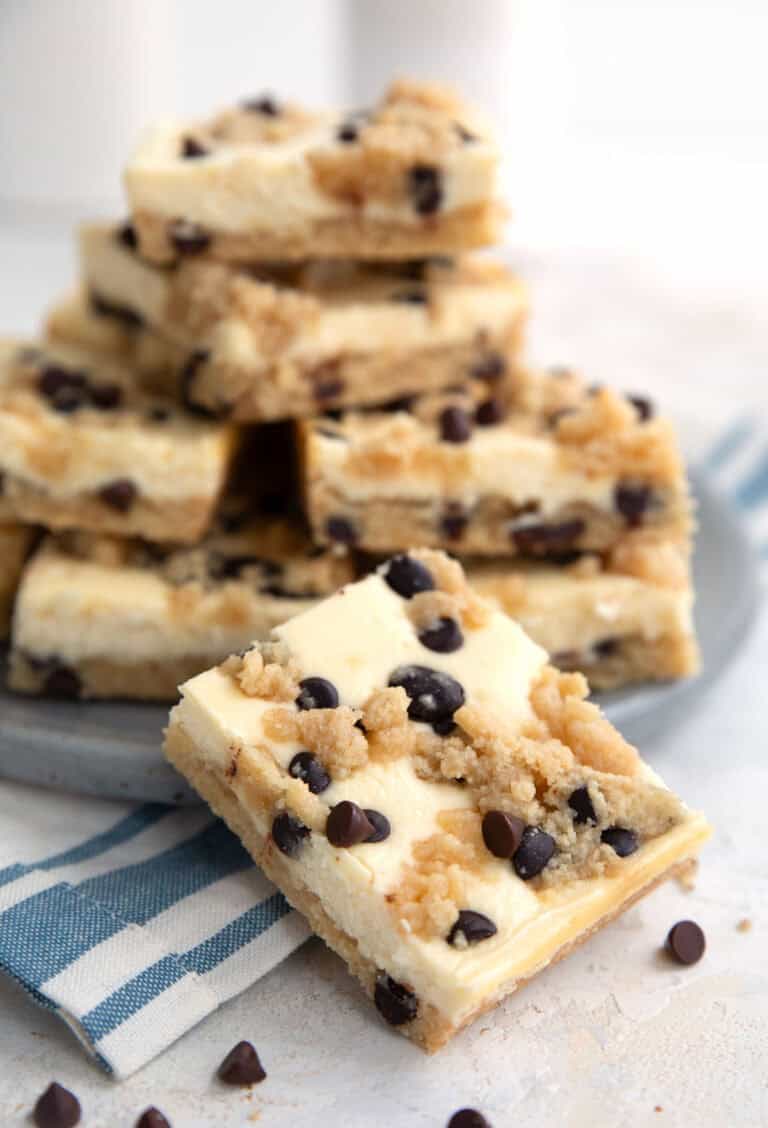 A Keto Chocolate Chip Cookie Cheesecake Bar sits in front a plate stacked with more bars.