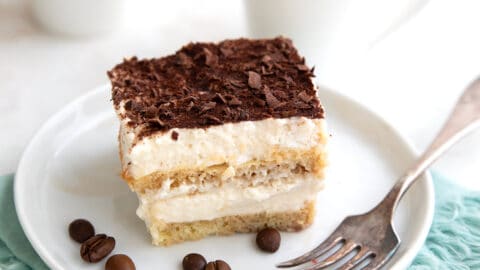 A slice of keto tiramisu on a white plate with cups of espresso in the background.