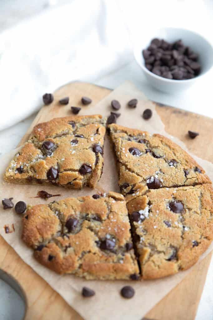 A large keto chocolate chip cookie cut into four pieces, sitting on a wooden cutting board with chocolate chips strewn around.