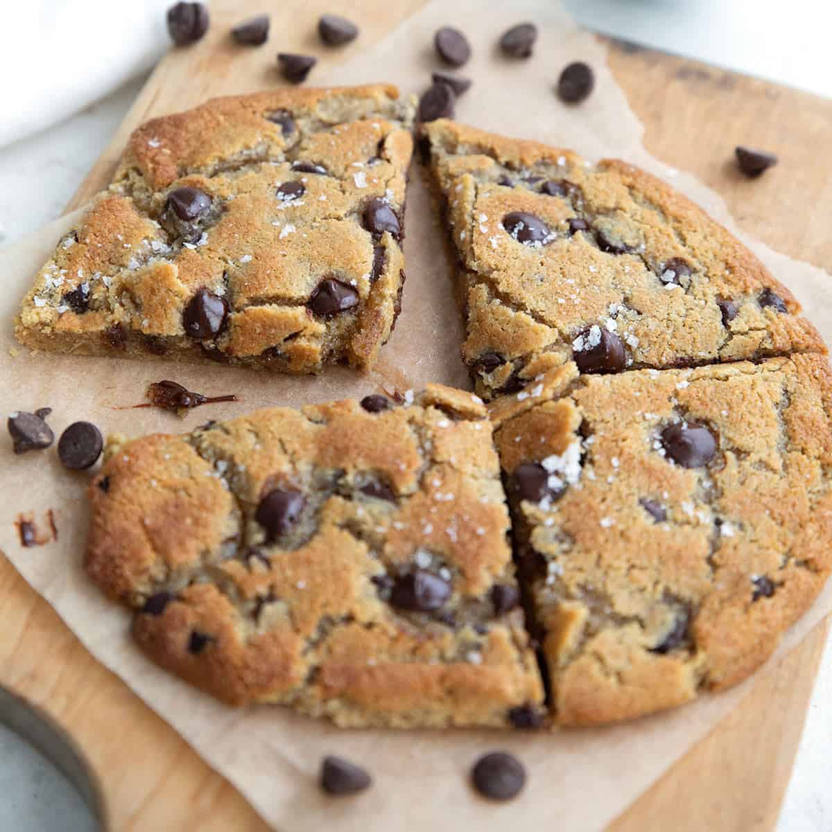 A large keto chocolate chip cookie cut into four pieces, sitting on a wooden cutting board with chocolate chips strewn around.