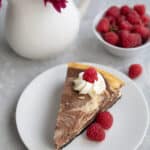 A slice of keto marble cheesecake on a white plate with raspberries and a vast of purple flowers.