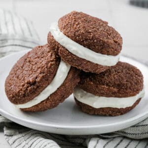 Three Keto Gingerbread Whoopie Pies piled up on a white plate over a striped napkin.