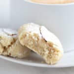 Close up shot of two keto amaretti cookies on a white plate with a cup of coffee, with a bite taken out of one of the cookies.