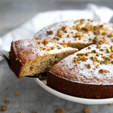 A cake lifter removing a piece of keto pistachio cake from a white cake stand.