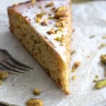 A slice of Keto Pistachio Cake on a gold plate lined with white parchment paper, with nuts strewn around.
